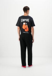 Young man facing away wears black t-shirt with orange boxing gloves and "Empire Fight Club" text, black pants, and red sneakers.