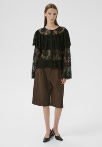 Sort blonde bluse med tierede flæser og lange ærmer, parret med brune shorts. Model bærer sorte hæle. Enkel baggrund.