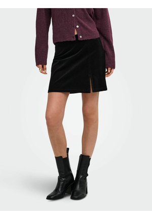 Femme portant une mini-jupe noire avec une fente à l'avant, un pull violet boutonné et des bottines noires à talons hauts, debout sur fond blanc.
