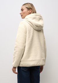 Chaqueta de forro polar crema con textura suave, capucha y dobladillo liso beige en contraste. Corte holgado y mangas largas. Jeans de mezclilla oscuros visibles.