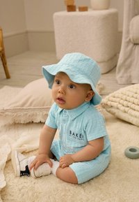 Mono corto azul claro con patrón y cuello, que presenta un logo. Combinado con un sombrero de cubo a juego y zapatos blancos, sentado sobre una superficie texturizada.