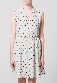 Abito senza maniche in tessuto crema chiaro con motivo a cuori neri, scollatura a V e vita elastica per una vestibilità regolabile.
