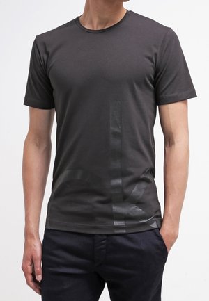 Mann trägt ein eng anliegendes dunkelgraues Kurzarm-T-Shirt mit dezentem vertikalem und diagonalem Streifendesign auf der Vorderseite sowie eine dunkle Hose.