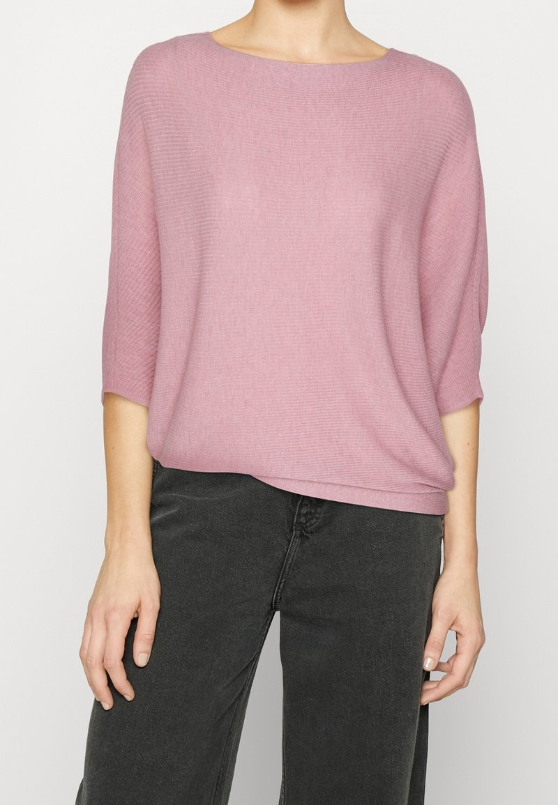 Pull en tricot côtelé rose clair avec un col large et une coupe ample. Manches longues et ourlet légèrement cintré. Associé à un pantalon large gris foncé.