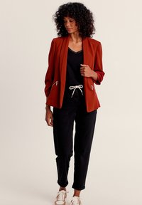 Rostfarbener Blazer mit strukturierten Schultern, vorderen Reißverschlusstaschen, Zweiknopfschluss, kombiniert mit schwarzen Hosen und Sneakers. Strukturierter Stoff.