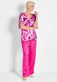 Blumenbluse in Rosatönen mit V-Ausschnitt und kurzen Ärmeln, kombiniert mit glänzenden, gerade geschnittenen rosa Hosen. Sandalen runden das Outfit ab.