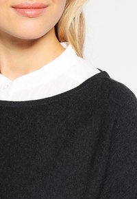 Maglione nero lavorato a maglia con un ampio scollo, indossato sopra una camicia bianca con bottoni caratterizzata da una trama trasparente e un colletto arrotondato.