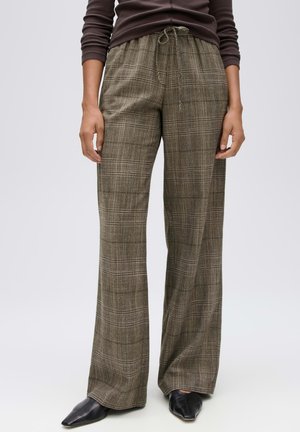 Personne portant un pantalon large à carreaux marron avec une taille à cordon et des chaussures noires à bouts pointus, debout devant un fond uni.