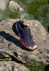 Fialové trailové běžecké boty s černými tkaničkami a oranžovými akcenty. Mají texturovanou podrážku a tvarovaný design pro podporu.
