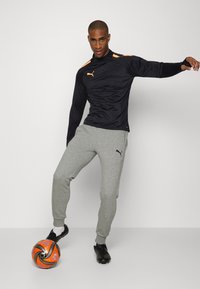 Schwarzes sportliches Zip-Up-Oberteil mit orangefarbenen Akzenten, kombiniert mit grauen Jogginghosen. Das Model steht mit einem Fußball auf schwarzen Fußballschuhen.