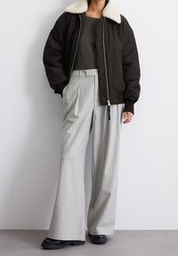 Svart bomberjacka med en krämfärgad fårskinnskrage, i kombination med ljusgrå rutig vida byxor och kraftiga svarta skor.