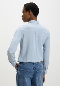 Persona con capelli ricci corti che indossa una camicia a maniche lunghe azzurro chiaro infilata in un paio di jeans blu a vita alta, in piedi contro uno sfondo neutro.