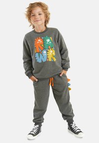 Grauer Sweatshirt mit bunten Monsterdesigns, kombiniert mit passenden grauen Sweatpants, die eine orangefarbene Kordel und verspielte bunte Akzente aufweisen.