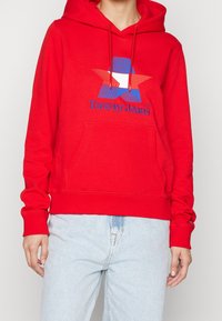 Felpa rossa con cappuccio e tasca a marsupio, caratterizzata da una grafica di stelle blu e bianche e testo "Tommy Jeans" ricamato in blu.