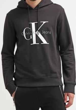 Person trägt einen schwarzen Calvin Klein Jeans Hoodie mit großem weißem "cK" Logo und hat die Hände teilweise in den Hosentaschen mit Reißverschlüssen.