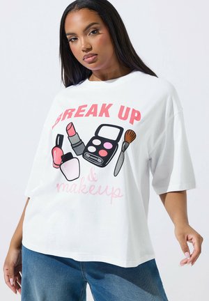 Kobieta ubrana w białą, oversize'ową koszulkę z napisem "BREAK UP & makeup" oraz ilustracjami kosmetyków, w połączeniu z niebieskimi dżinsami.