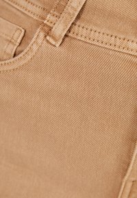 Vaaleanruskea denim-kangas, jossa on hieno tekstuuri, näkyvät saumat ja sileä pinta. Lähikuva materiaalin yksityiskohdista.