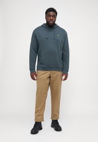 Hoodie verde oscuro con un bolsillo frontal y cordones, combinado con pantalones marrón claro y botas negras con cordones. Diseño sencillo, sin estampados.