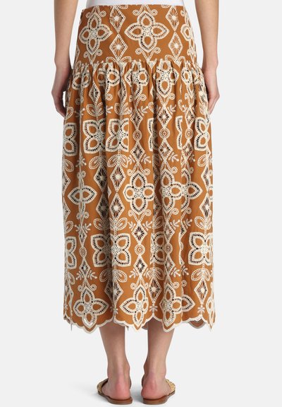 Fustă midi cu broderie florală albă din dantelă pe țesătură maro, tiv ondulat, purtată cu sandale plate, văzută din spate.