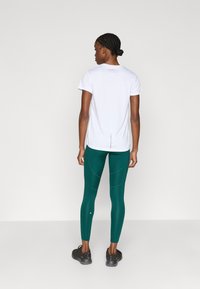 Balti sportiniai marškinėliai ir žalsvos spalvos tamprės su siūlių akcentais, derinamos su juodais sportiniais bateliais. Lygus audinys ir aptemptas dizainas.