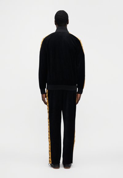 Μαύρο βελούδινο φόρμα που π εριλαμβάνει ένα μπουφάν με φερμουάρ και ψηλό γιακά, καθώς και κίτρινες ρίγες με λεοπάρ σχέδιο στα πλάγια, σετ με παντελόνι που ταιριάζει.