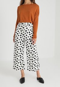 Rostfärgad långärmad topp med avslappnad passform, parat med vita vida byxor med svarta prickar. Svarta slip-on-skor fulländar looken.