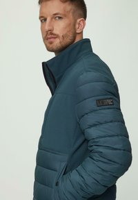 Teal pufferjack met een hoge kraag; heeft een mix van gladde en geborduurde texturen. Bevat een met rits afsluitbare zak en een merkpatch op de arm.