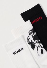 Trzy pary skarpet: czarne z prążkowanej tkaniny z czerwonym logo, białe z prążkowanej tkaniny z czarnym logo i grafiką oraz dodatkowy szczegół graficzny.