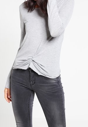 Maglietta grigia a maniche lunghe con dettaglio a nodo torsadato sul davanti, realizzata in un tessuto morbido, abbinata a jeans slim-fit grigi scuri.