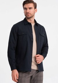 Camisa de botones azul marino oscuro con dos bolsillos en el pecho, mangas largas y un ajuste casual, superpuesta sobre una camiseta beis clara.