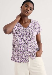 Top floral morado con mangas cortas, cuello en V y detalles de botones. Presenta patrones florales en blanco y negro sobre una tela suave. Combinado con pantalones claros.
