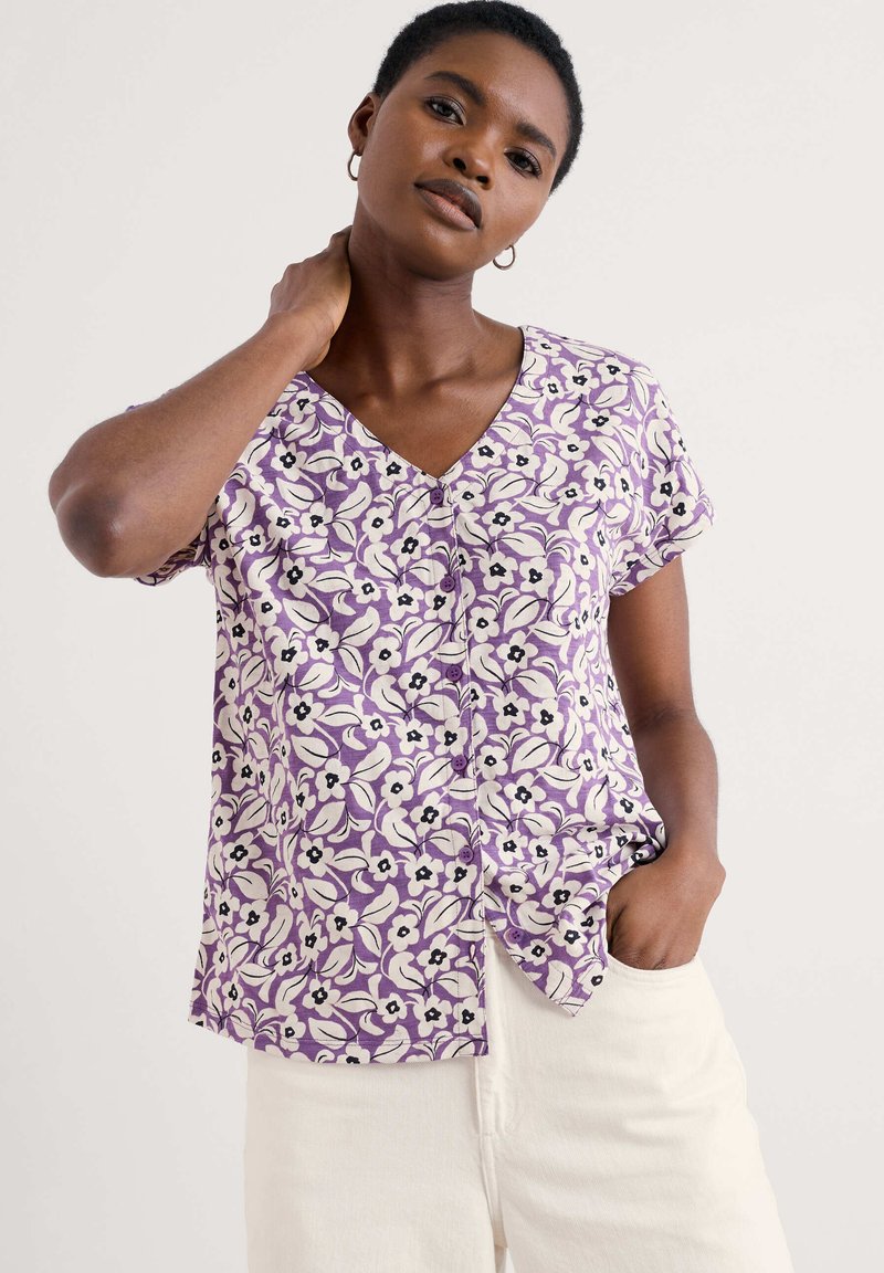 Top floral morado con mangas cortas, cuello en V y detalles de botones. Presenta patrones florales en blanco y negro sobre una tela suave. Combinado con pantalones claros.