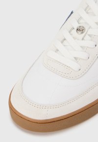 Bijela tenisica od glatke kože s okomitim akcentima od antilopa, okruglim vrhom, ravnim bijelim vezicama i svijetlo smeđim gumenim potplatom.