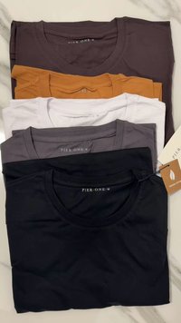 Quattro t-shirt piegate di colori diversi: grigio scuro, giallo senape, bianco e nero. Realizzate in cotone biologico con colli rotondi.