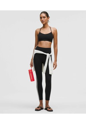 Frau in schwarzem Sport-BH und Leggings mit weißen Seitenstreifen, weißem Sweatshirt, das in der Taille gebunden ist, hält eine rote Lululemon-Wasserflasche.