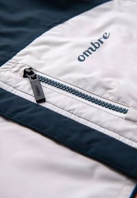 Leichte, weiße Nylonjacke mit einem dunkelblauen Akzent, ausgestattet mit einer zipbaren Tasche und einem "Ombre"-Logo in Marineblau. Glatte Textur mit kontrastierenden Farben.