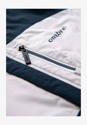 Lightweight, white nylon jacket with a dark blue accent, featuring a zippered pocket and "ombre" logo in navy. Smooth texture with contrasting colors.