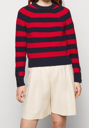 Femme portant un pull en tricot rayé rouge et bleu marine avec des détails de boutons sur les manches et un short large couleur crème.