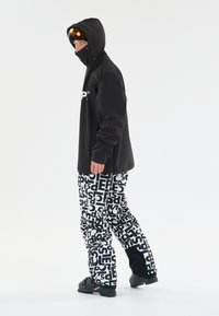 Sort hættejakke med logo foran, parret med oversized bukser med sort og hvidt mønster. Footwear præsenterer et tekstureret design.