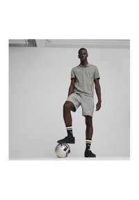 Graues Poloshirt und Shorts aus weichem Stoff, mit einem dezenten Logo. Schwarze Sneaker und gestreifte Socken. Ball am Fuß. Schlichter Hintergrund.