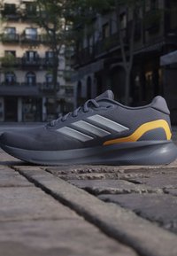 Chaussure de course gris foncé avec des bandes argentées et un accent jaune au talon, posée sur une rue pavée avec un arrière-plan urbain flou.