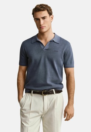 Man in een blauw poloshirt met korte mouwen, ingestopt in beige broek met een gevlochten bruine riem, handen in de zakken.