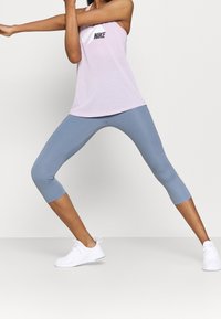 Nike-vaaleanpunainen hihaton paita, jossa on musta logo, yhdistettynä sinisiin capri-leggingseihin ja valkoisiin urheilujalkineisiin. Varustettu tiukalla muotoilulla ja pehmeällä tekstuurilla.