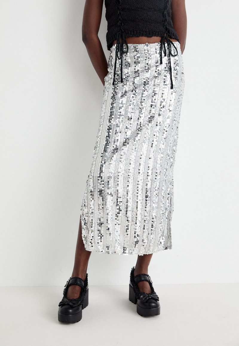 Jupe midi en sequins argentés avec des rayures verticales, une fente sur le côté et une texture lisse. Associée à des chaussures à plateforme noires et épaisses.