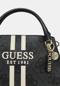 Sac à main noir avec des rayures blanches, le logo "GUESS" et un porte-clés avec le nom de la marque et l'année de création, "EST 1981."