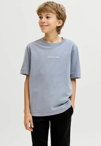 Αγόρι με κοντά ανοιχτό καστανά μαλλιά φοράει γαλάζιο T-shirt με το κείμενο «Originals Studio» και μαύρο παντελόνι, στέκεται με το ένα χέρι στην τσέπη.