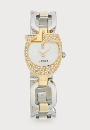 Montre en or avec un large cadran circulaire orné de cristaux. Bracelet en métal argenté et doré avec un design épais. Nom de la marque visible.