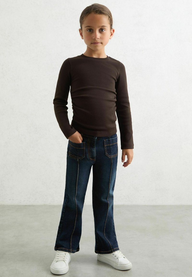 Brun langærmet skjorte, mørkeblå flagrende jeans med orange syninger og hvide sneakers, stående på et lyst gråt gulv foran en tekstureret baggrund.