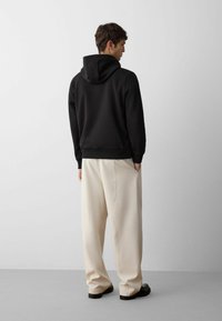 Felpa nera con cappuccio e finitura testurizzata, abbinata a pantaloni beige chiaro e larghi, e scarpe nere. Design semplice, dettagli minimi.