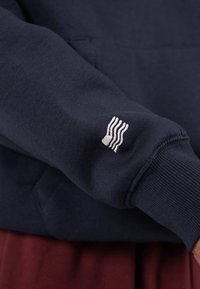 Granatowy sweatshirt z prążkowanymi mankietami, z małym białym logo w faliste linie na lewym rękawie. Miękka tekstura z mieszanki bawełny.